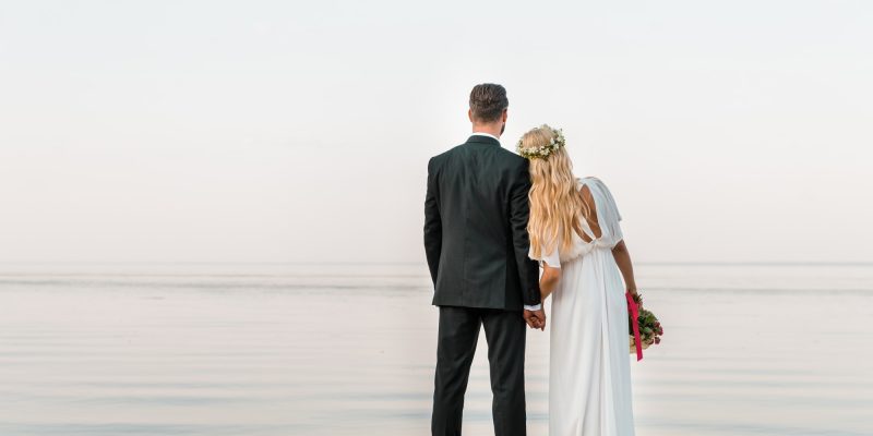 back view of wedding couple standing on beach with wedding bouquet and looking at sea